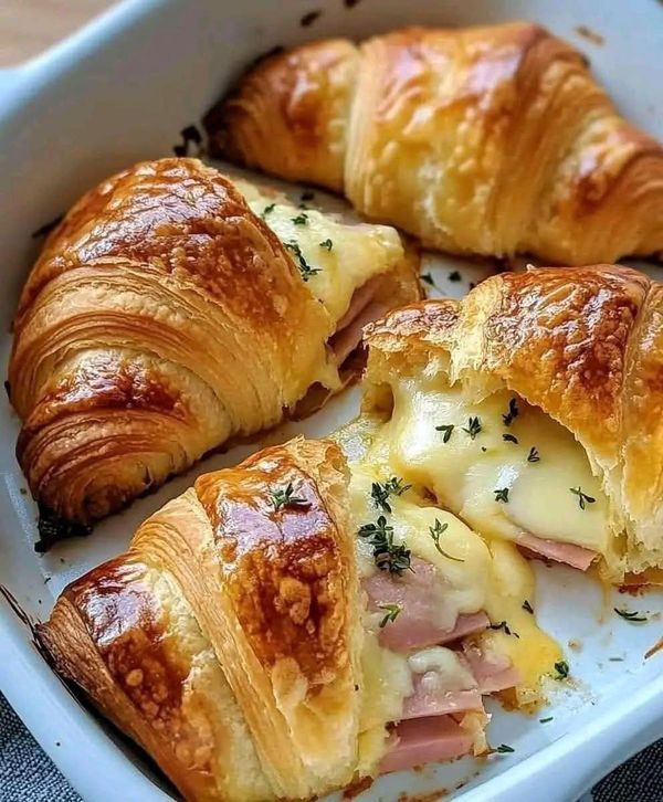 Delicious ham and cheese croissants with melted cheese, topped with herbs, in a white baking dish.