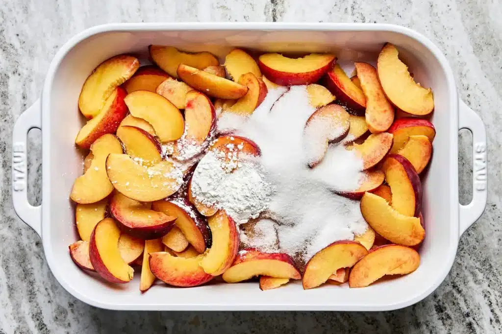 Sliced peaches with sugar and flour in a baking dish, ready for cobbler preparation.