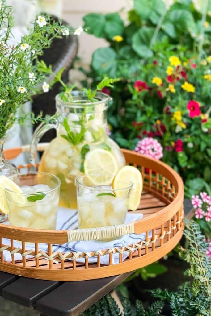 Refreshing lemonade with lemon slices on a wicker tray outdoors, surrounded by vibrant flowers and greenery.