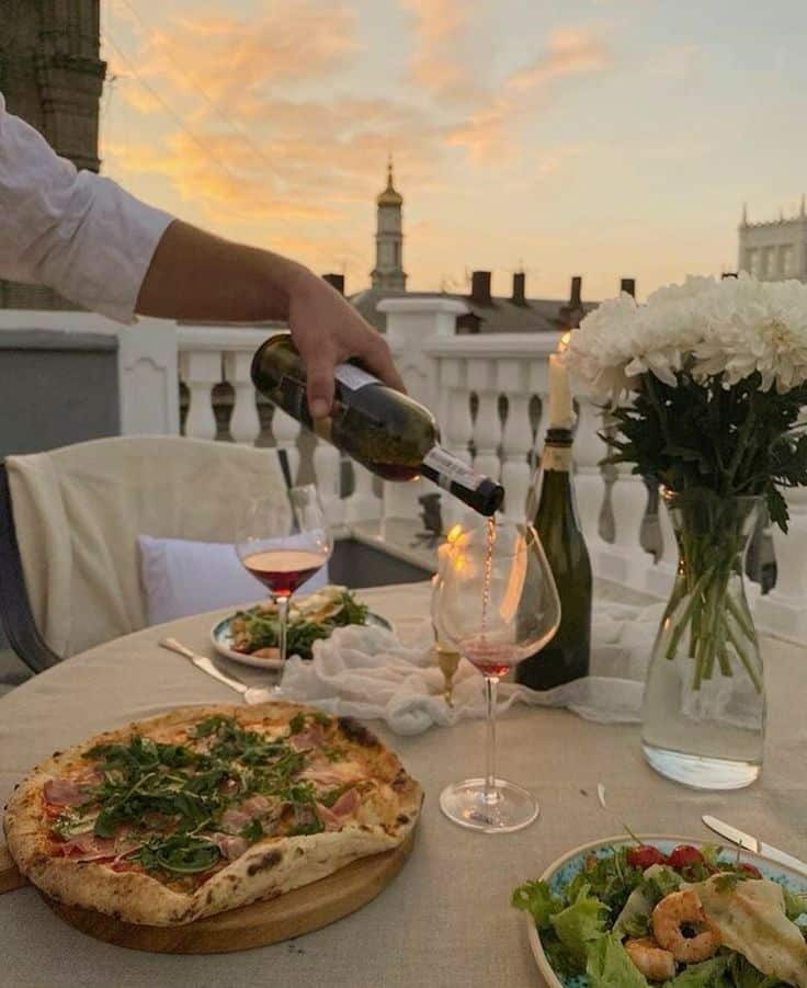Outdoor dining with pizza, salad, and wine at sunset, elegant rooftop setting with flowers.