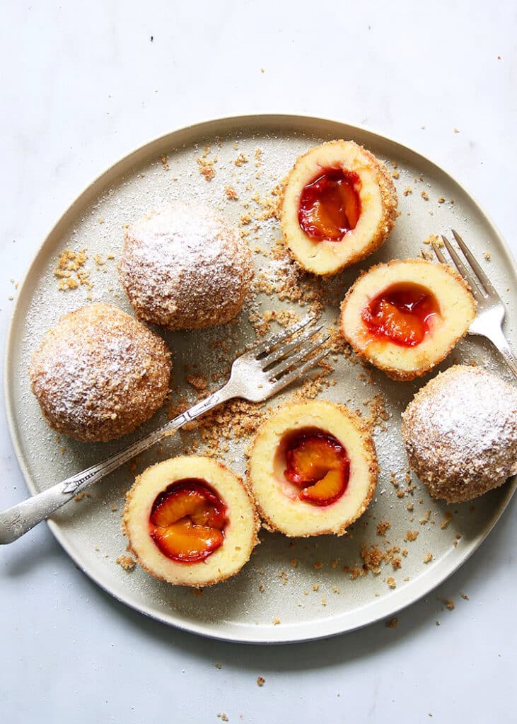 Plum dumplings dusted with powdered sugar on a plate, with crumbs and forks, showcasing a delicious dessert presentation.