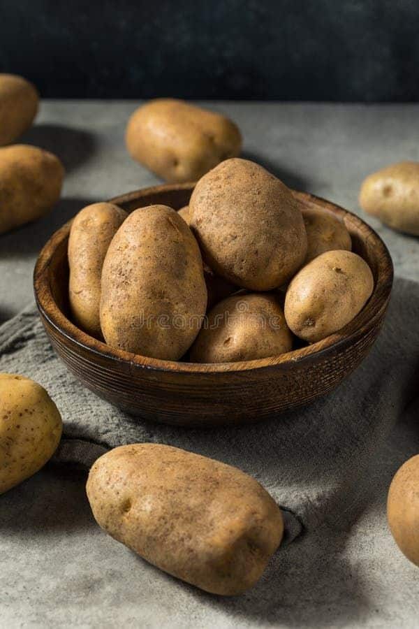 Wooden bowl filled with fresh brown potatoes on a textured cloth, surrounded by more potatoes on a gray surface.