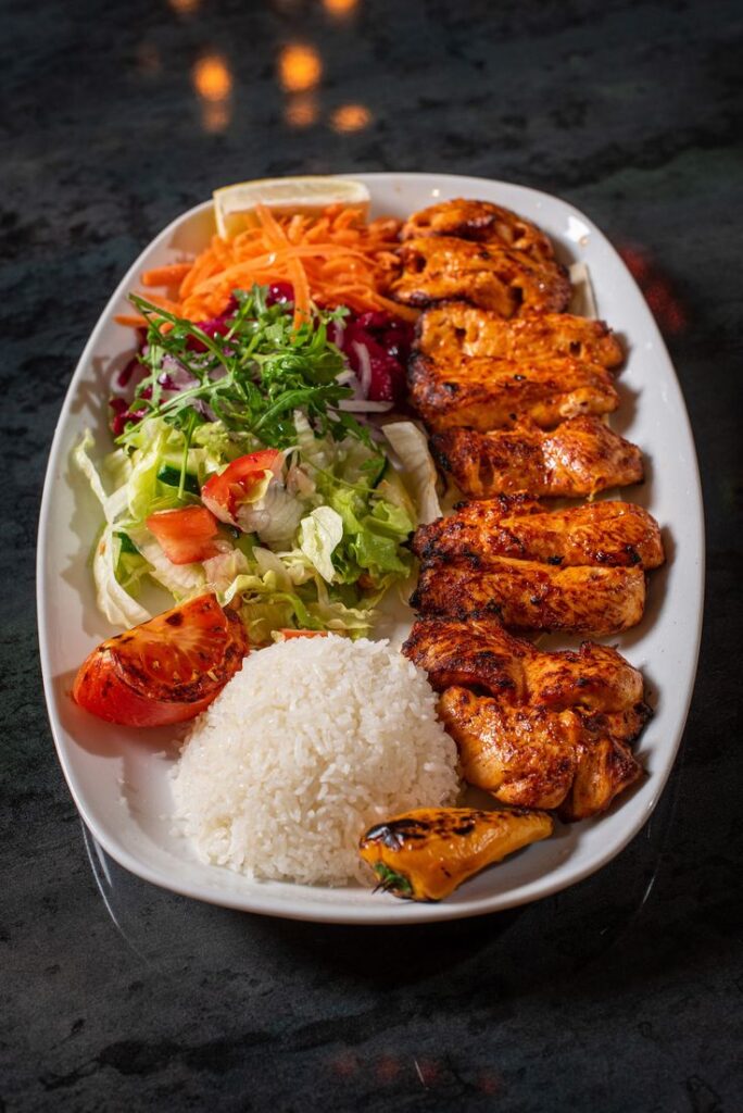 Grilled chicken platter with rice, fresh salad, roasted vegetables on a white plate, served on a dark table.