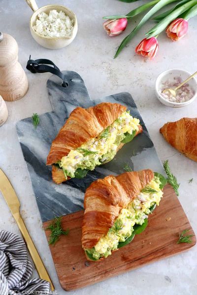 Fresh croissant sandwiches with egg salad and dill on a marble board, surrounded by tulips and tableware.
