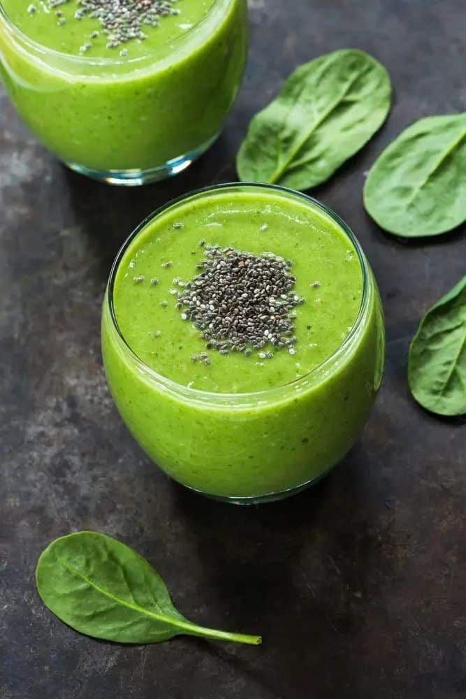 Green smoothie with chia seeds in a glass, surrounded by fresh spinach leaves on a rustic surface.