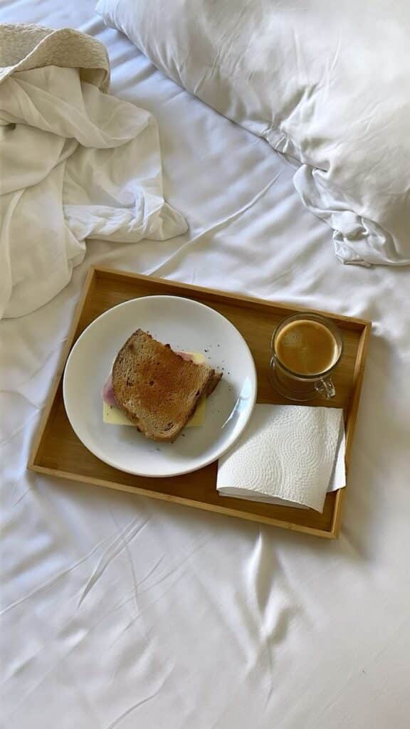 Breakfast in bed with a toasted sandwich and espresso on a wooden tray with white linens.