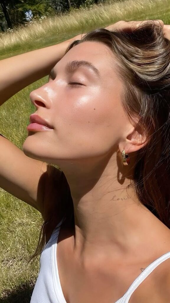 Woman enjoying sunlight in a field, eyes closed, relaxed expression, wearing hoop earrings and a white top.