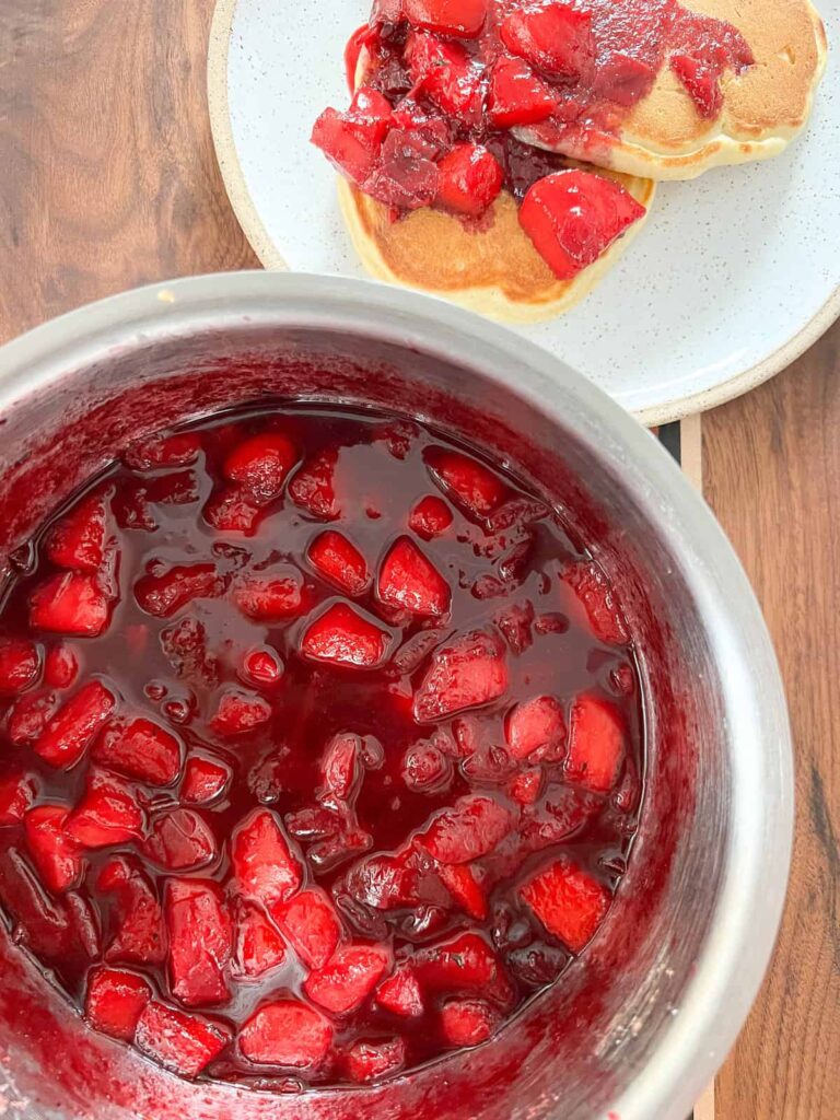 Homemade strawberry sauce in a bowl with pancakes on a plate, topped with bright red, chunky strawberry syrup.