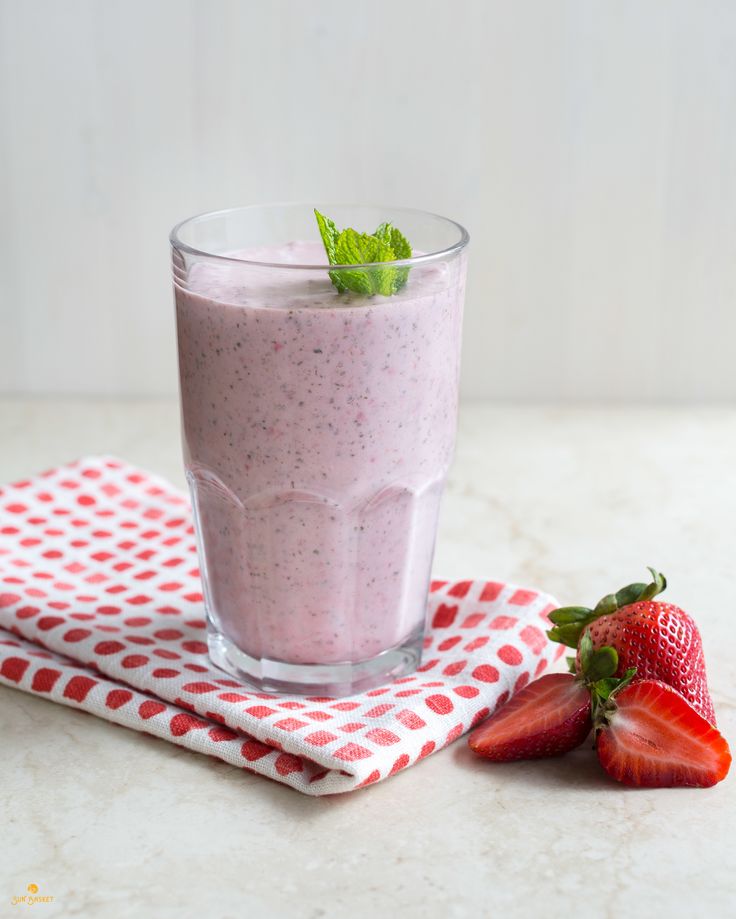 Strawberry smoothie in a glass with mint garnish on a polka dot cloth, next to fresh strawberries on a marble surface.