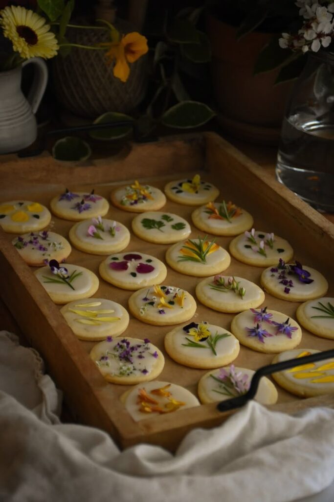 Wooden tray with decorated cookies topped with colorful edible flowers, surrounded by pots and a vase.