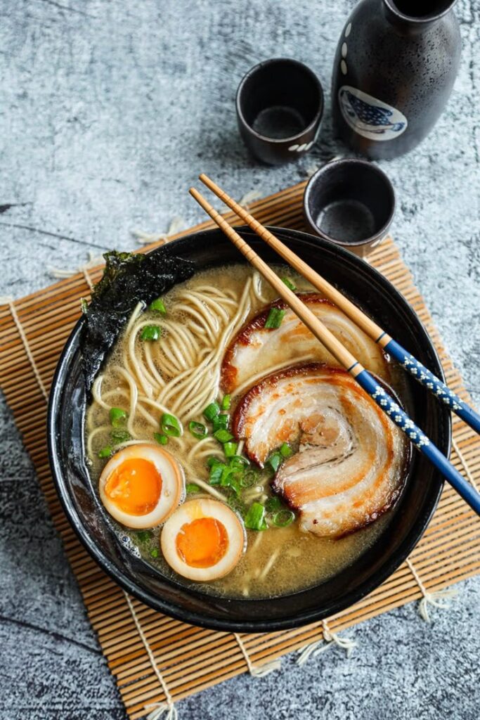 Delicious bowl of ramen with pork, eggs, and green onions, served with chopsticks and sake. Perfect Japanese meal.