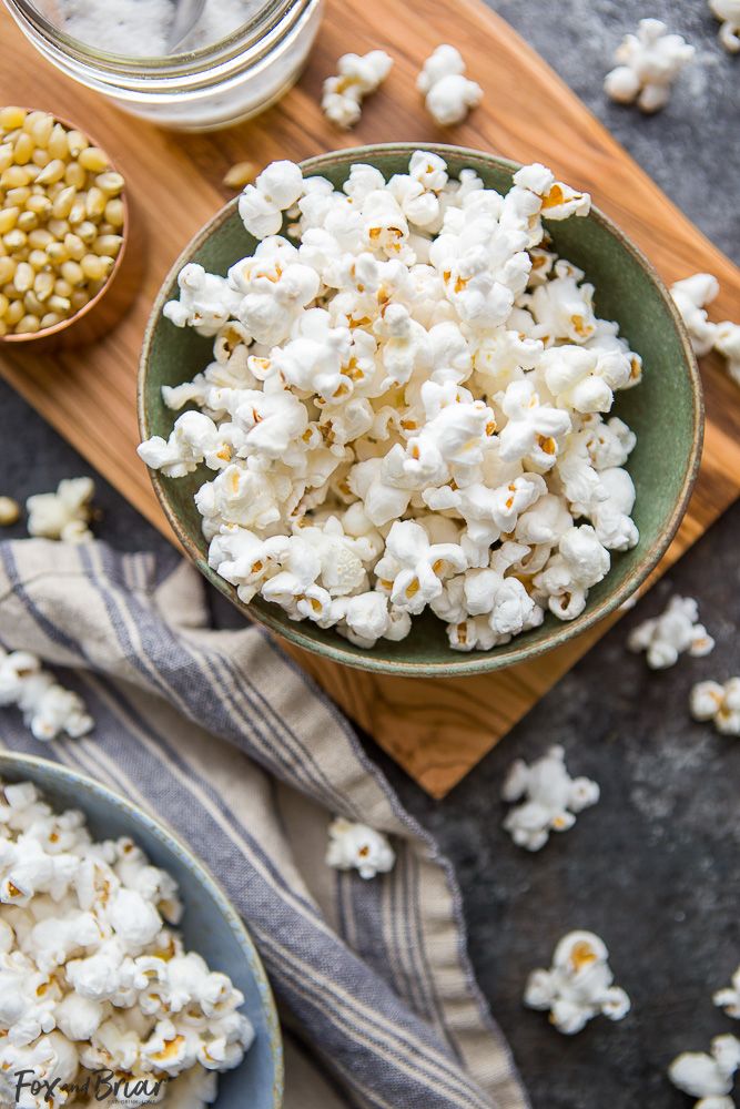 Bowl of fluffy, freshly popped popcorn next to kernels in a rustic kitchen setting on a wooden board.