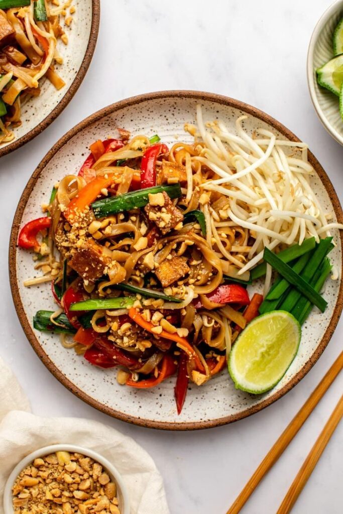 Delicious pad thai noodles with vegetables, garnished with crushed peanuts, lime, and bean sprouts on a plate.