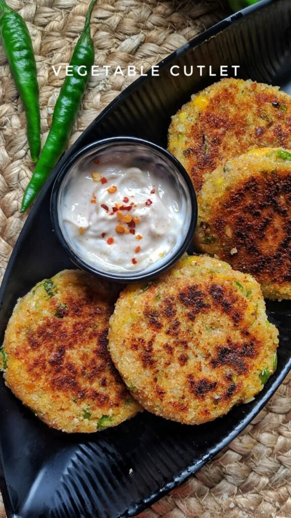 Crispy vegetable cutlets with creamy dip served on a black plate, garnished with chili flakes and green chilies.