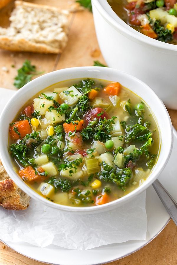 Hearty vegetable soup with carrots, peas, and kale served in a white bowl on a wooden table with bread.