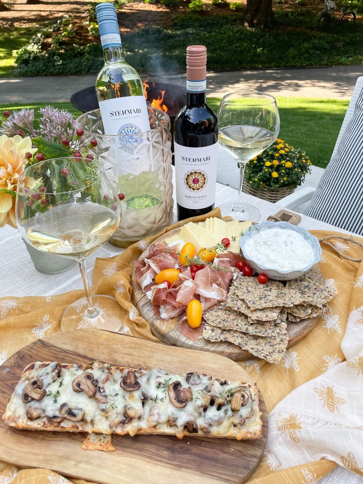 Outdoor picnic setup with wine, mushroom pizza, cheese, prosciutto, and crackers on a table adorned with flowers.