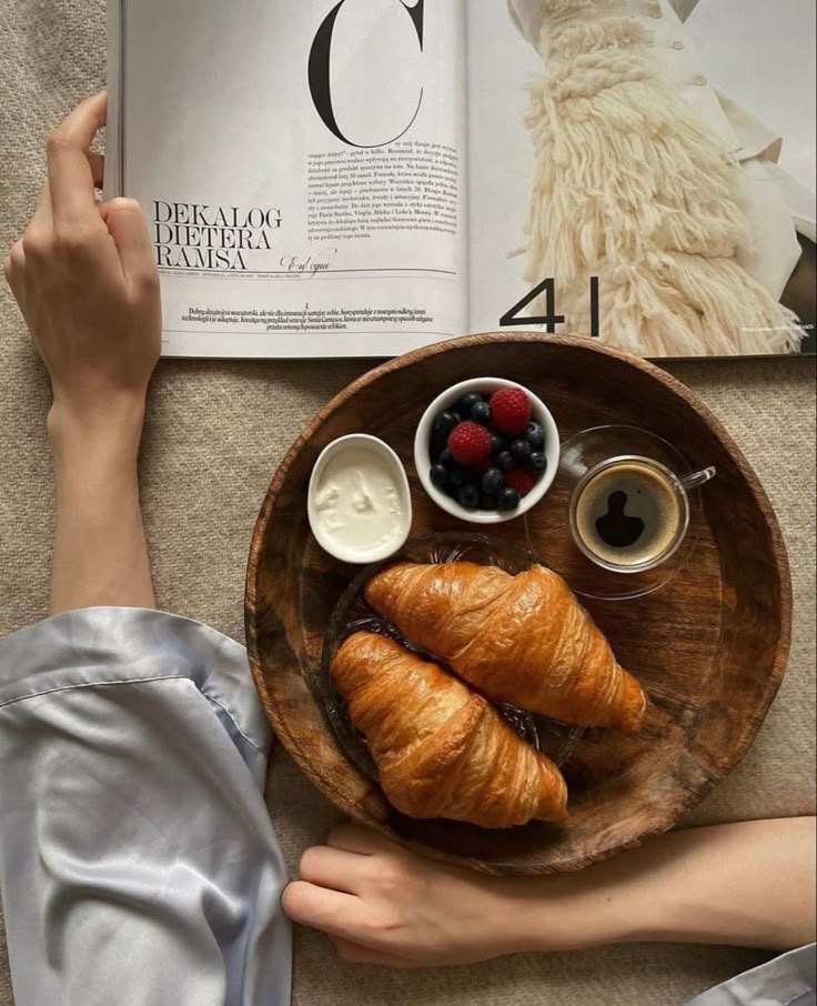 Reading a magazine in bed with a breakfast tray of croissants, berries, yogurt, and espresso. Cozy morning vibe.