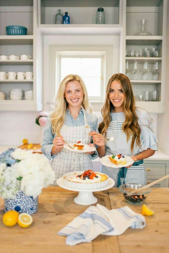 Two women enjoying homemade fruit cakes in a cozy kitchen setting with fresh flowers and citrus fruits on the table.