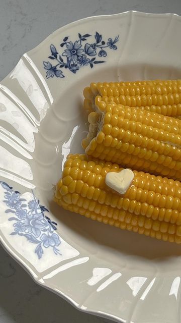 Three ears of corn with heart-shaped butter on a floral plate, highlighting freshness and homestyle cooking.