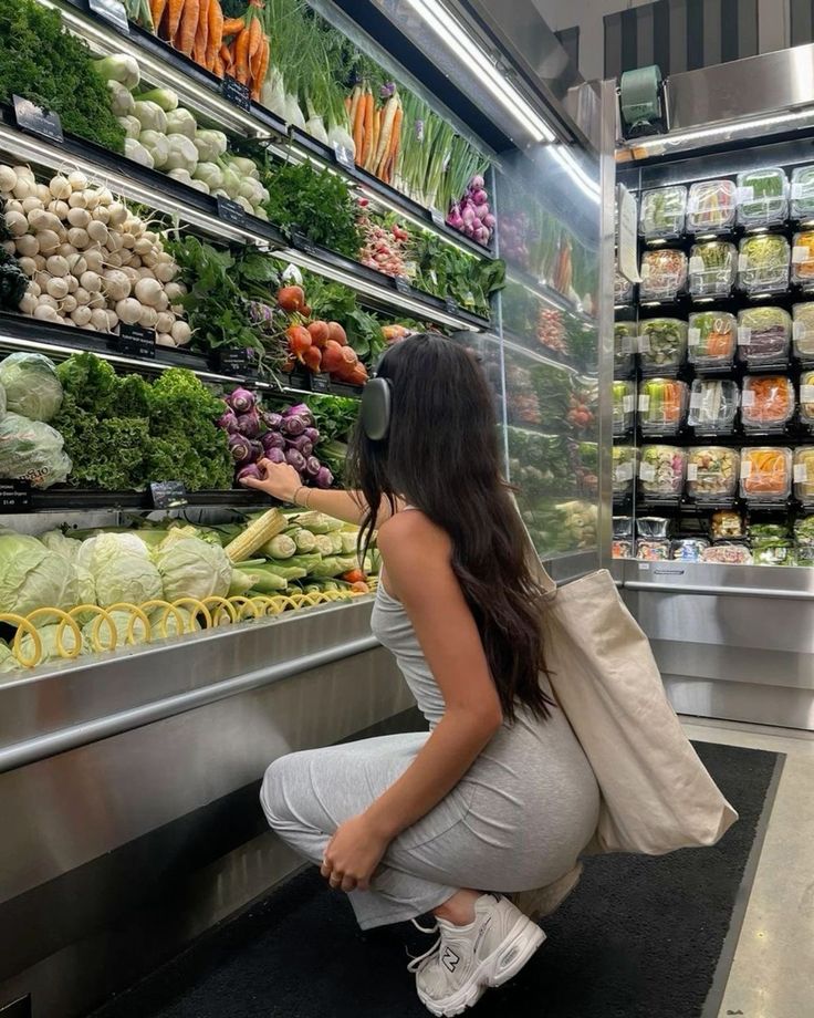Woman shopping for fresh vegetables at a grocery store, wearing headphones and holding a tote bag.