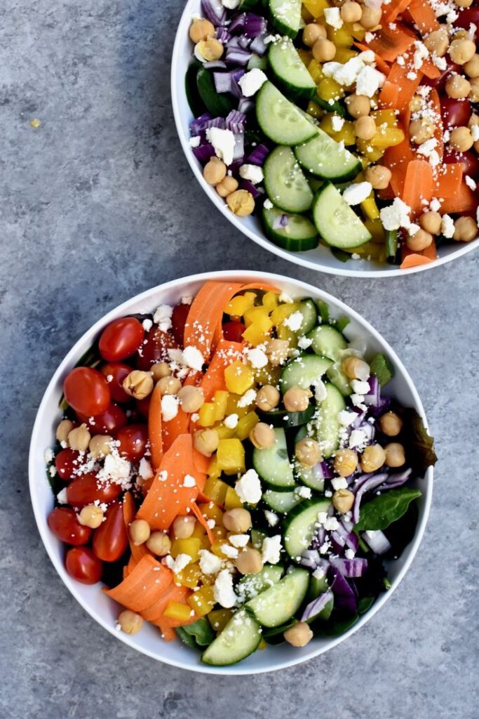 Close-up of colorful Mediterranean salad bowls with cucumbers, tomatoes, carrots, chickpeas, and feta on a stone background.