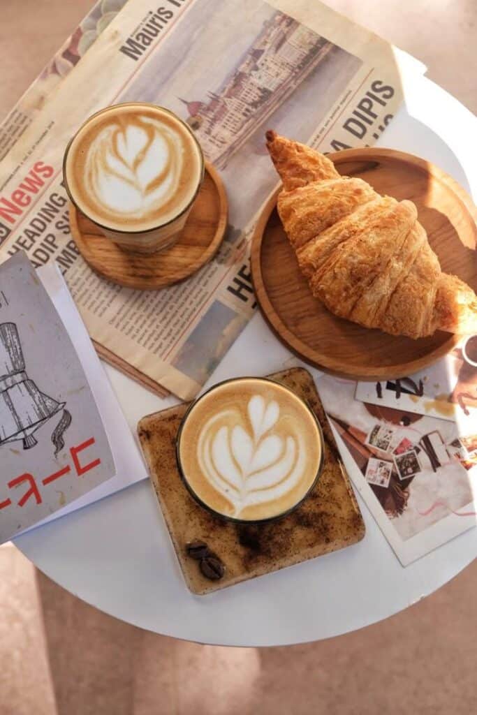 Two cappuccinos with latte art and a croissant on a table with newspapers, creating a cozy breakfast scene.