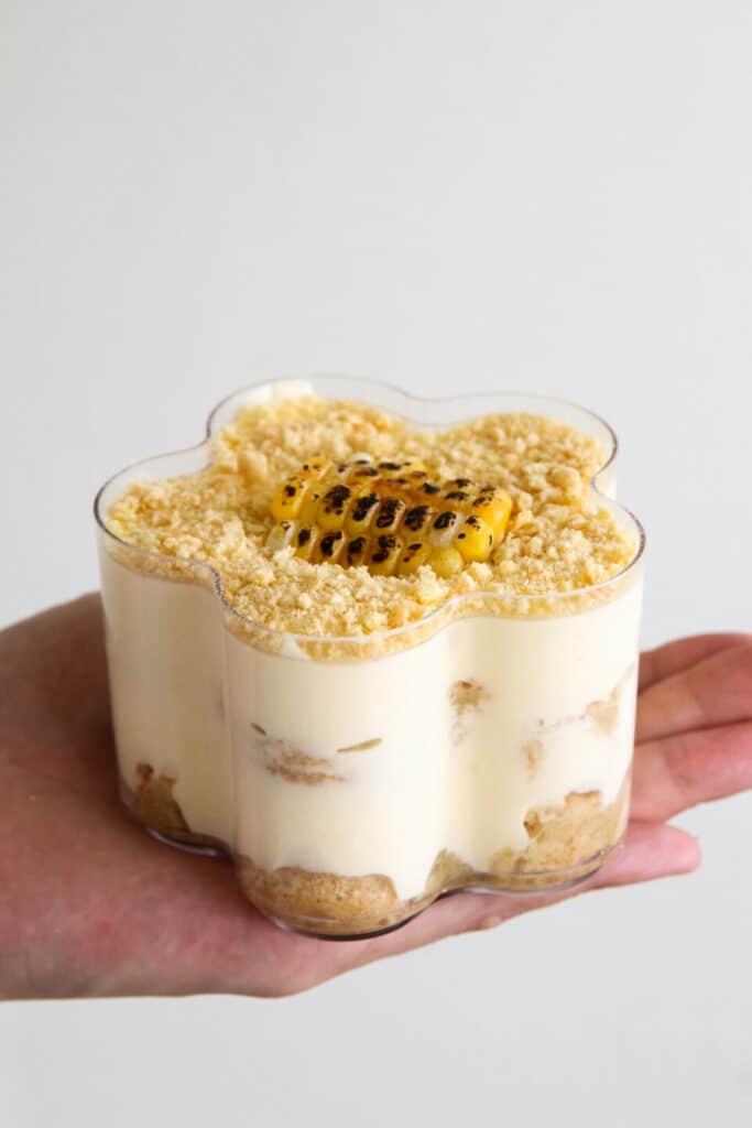 Creamy dessert in a flower-shaped cup, topped with grilled corn and crumbled biscuits, held in hand.