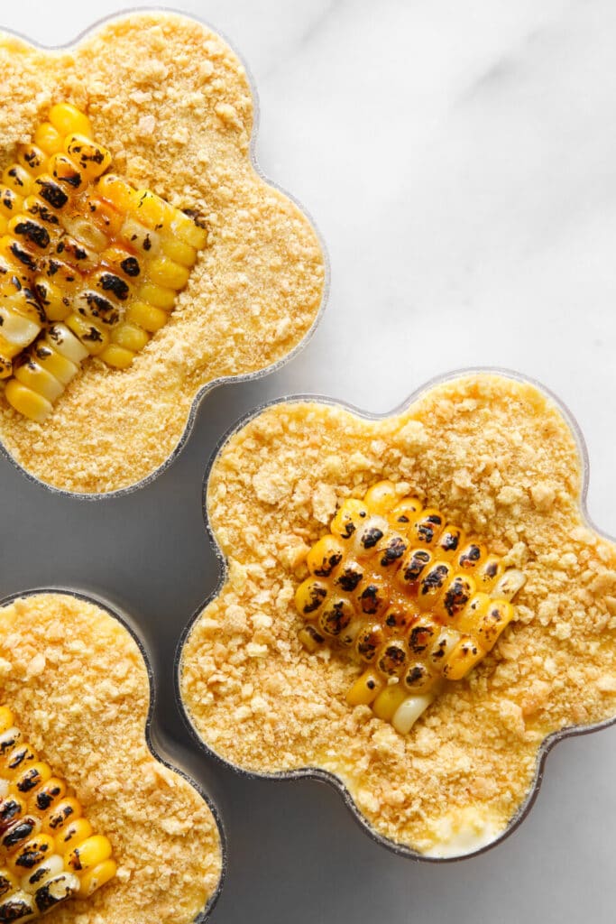 Three individual corn puddings topped with crushed crackers and grilled corn pieces on a light background.