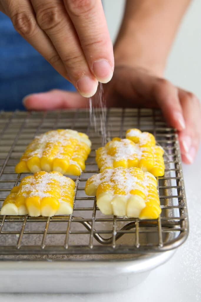 Sprinkling sugar over corn-shaped cookies on a cooling rack.