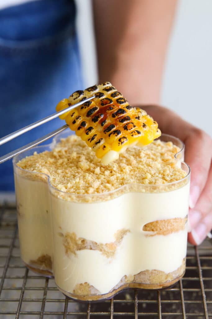 Grilled corn being placed on a creamy dessert topped with crumble, showcasing a mix of textures and flavors.