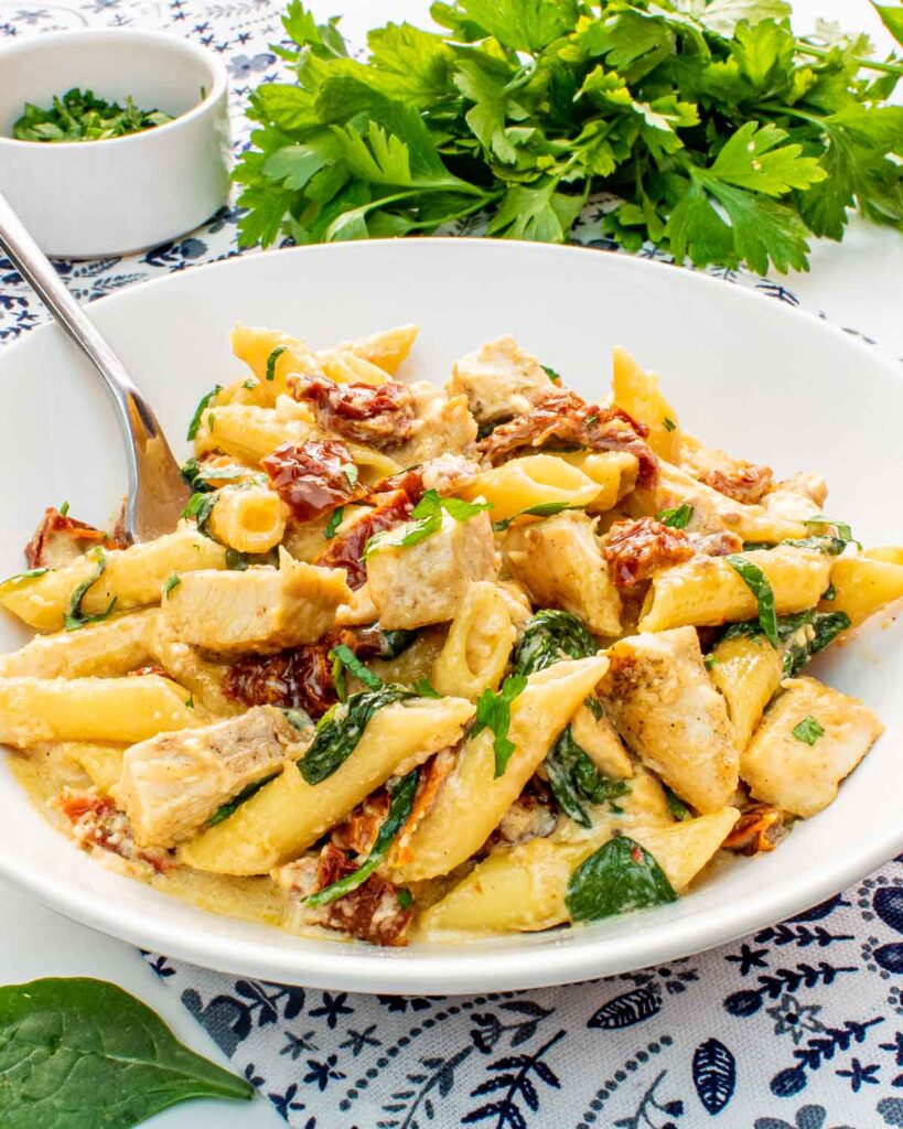 Creamy penne pasta with chicken, sun-dried tomatoes, and spinach, garnished with fresh herbs in a white bowl.