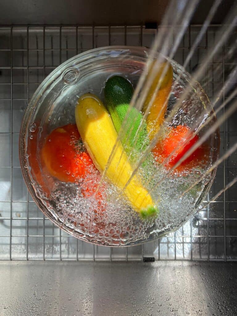 Washing colorful vegetables in a glass bowl under running water in a kitchen sink.