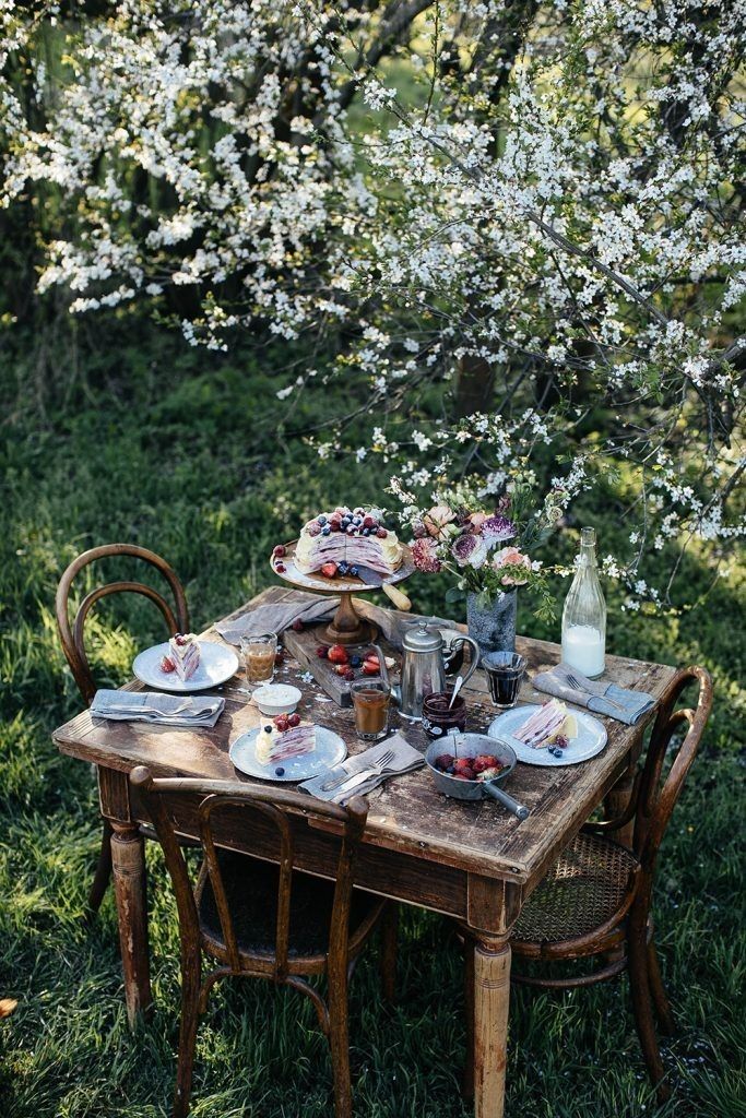 Rustic outdoor brunch setting with wooden table, pastries, flowers, and cherry blossom branches overhead.