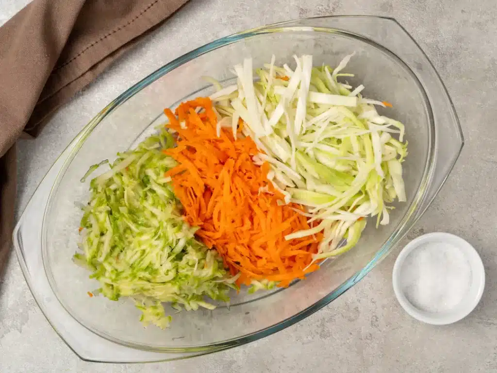 Grated carrots, cabbage, and zucchini in a glass bowl, with a dish of salt nearby, on a textured surface.