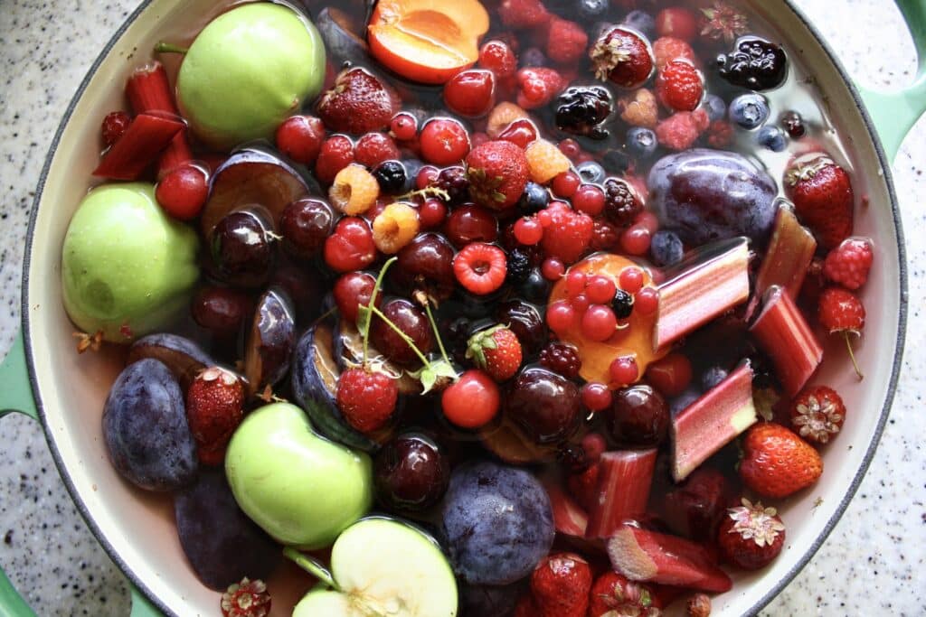 Fresh fruits and berries soaking in a pot, including apples and strawberries, for jam or preserves.