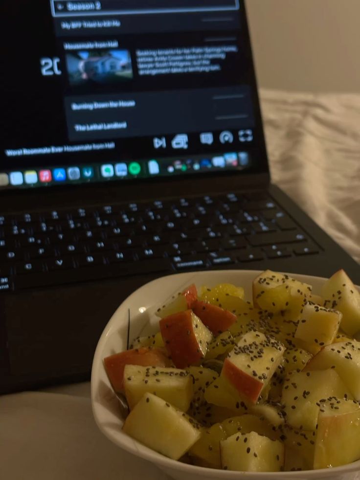 Bowl of fresh fruit salad beside laptop on bed, perfect for a cozy snack while watching movies or working online.