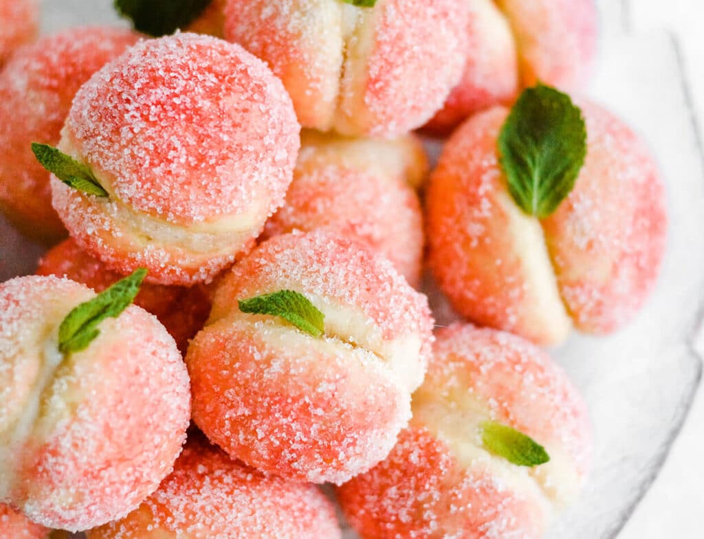 Sugary peach-shaped cookies with mint leaves, arranged in a pile on a white plate.