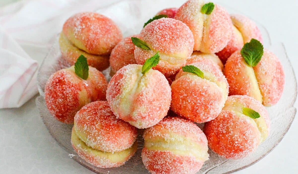 Sugar-coated peach-shaped cookies with mint leaves on a glass plate.