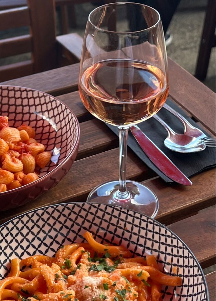 Pasta dishes and a glass of rosé wine on a wooden table, perfect for an al fresco Italian dining experience.