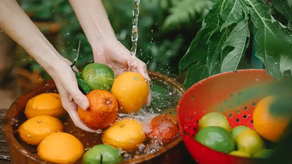 Hands washing fresh citrus fruits in a wooden bowl with water, surrounded by green leaves and a red colander.