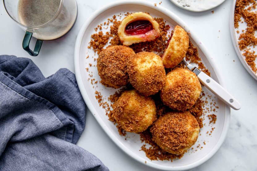 Plate of plum dumplings with breadcrumbs, a fork, and a cup of coffee on a marble surface.