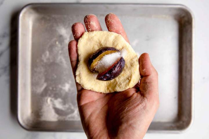 Hand holding dough with a plum and sugar inside, ready for baking on a tray.