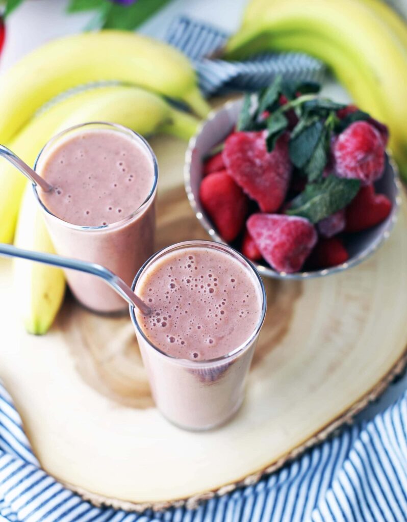 Two strawberry banana smoothies in glasses with metal straws, fresh bananas, and strawberries in the background.