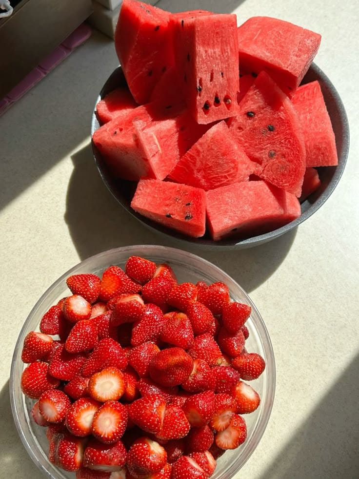 Bowls of fresh watermelon slices and strawberries on a sunny table, showcasing vibrant red summer fruits.