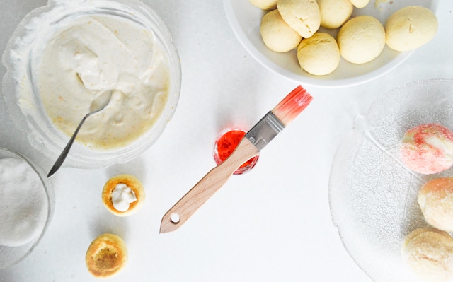 Table with baking ingredients: dough balls, cream, sugar, and a brush with red glaze, ready for pastry decoration.