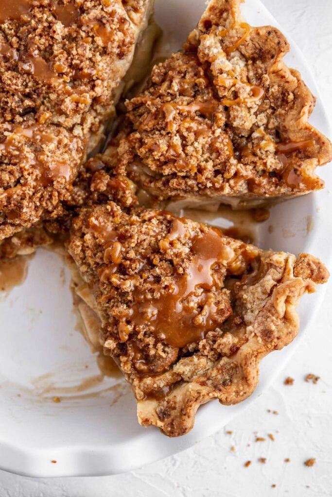 Three slices of caramel apple pie with crumb topping in a white dish, showcasing delicious textures and indulgent sweetness.