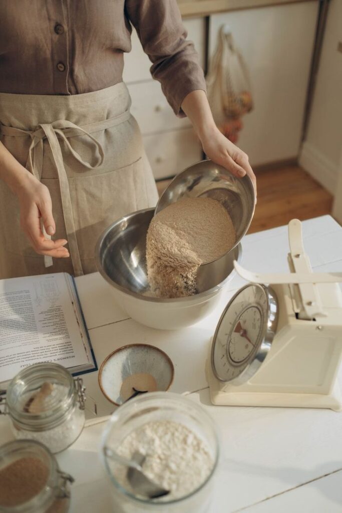 Person pouring flour into bowl while baking, surrounded by kitchen scales, cookbook, and ingredients.