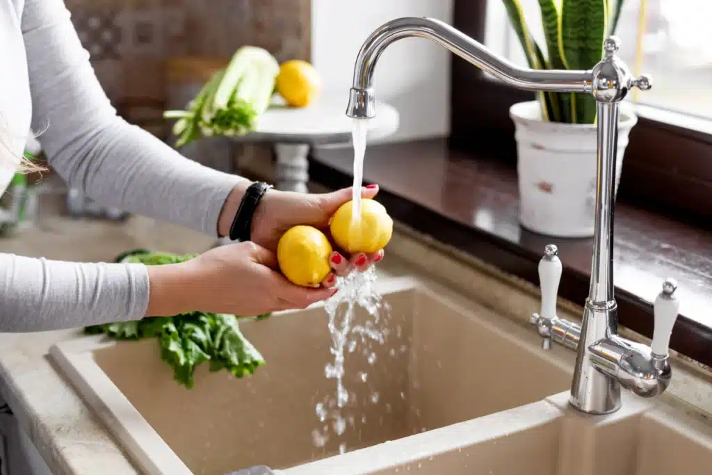 Washing fresh lemons under a kitchen faucet, with a green plant and vegetables in the background.