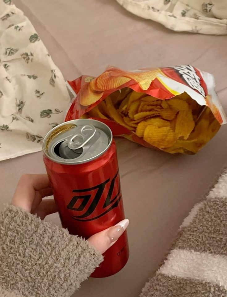 Hand holding a red soda can, with an open bag of wavy potato chips on a cozy blanket-covered bed.