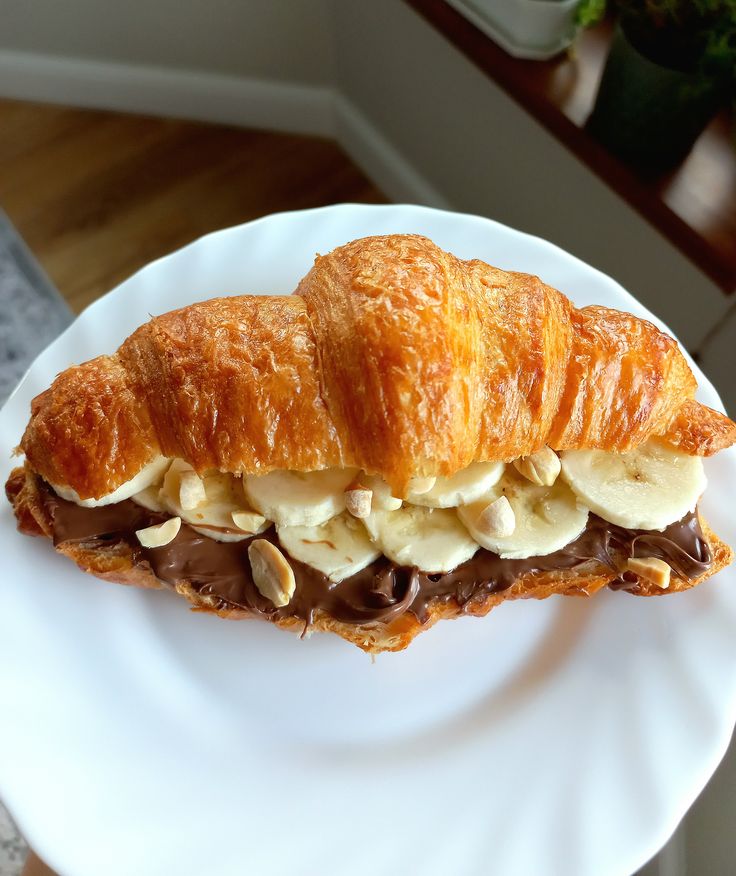 Golden croissant filled with chocolate spread, banana slices, and nuts on a white plate.
